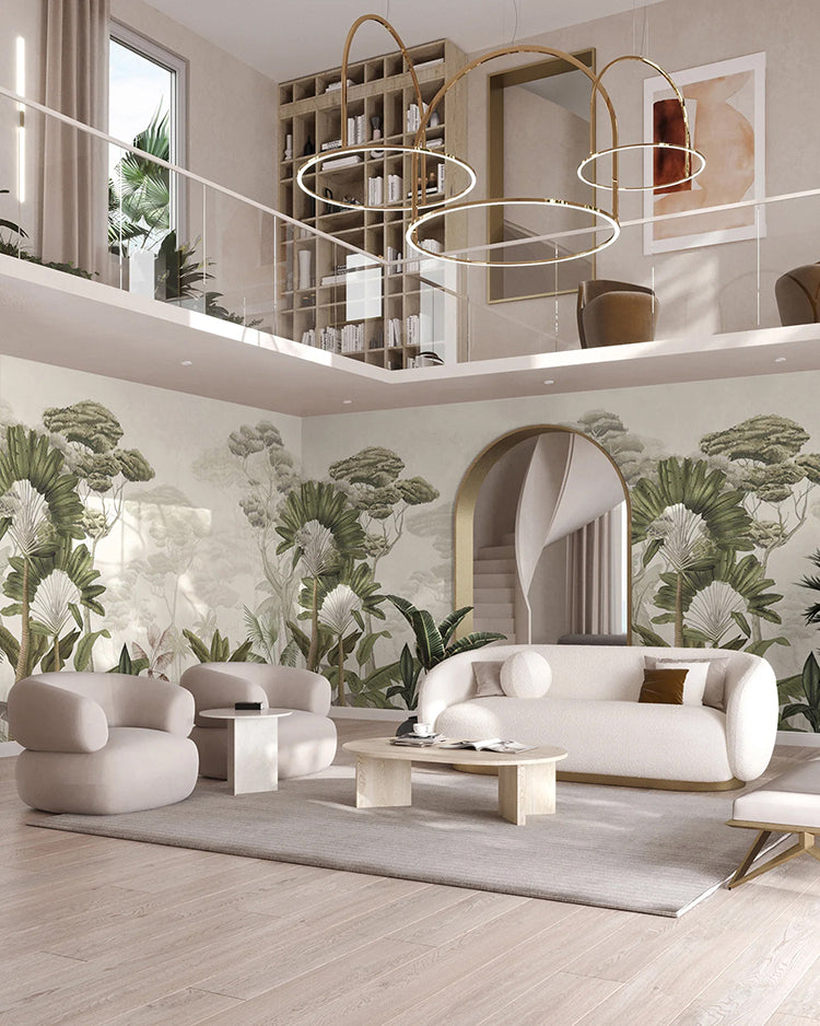 In this spacious, modern living room, natural light floods through tall windows, illuminating a two-level design. The main wall boasts the pale Garden Eden, Tropical Mural Wallpaper in Sand,  which transports you to a tropical paradise. The room is furnished with a beige sofa set, a wooden coffee table, and unique circular chandeliers, creating an inviting blend of contemporary aesthetics.