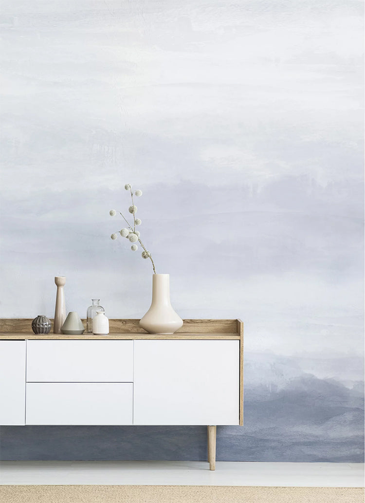 In a room, a wall features the Vista Watercolor Ombre Wallpaper in Dusky Blue, creating a striking contrast with a white table. The table is adorned with multiple ceramic vases, adding to the room’s aesthetic appeal.